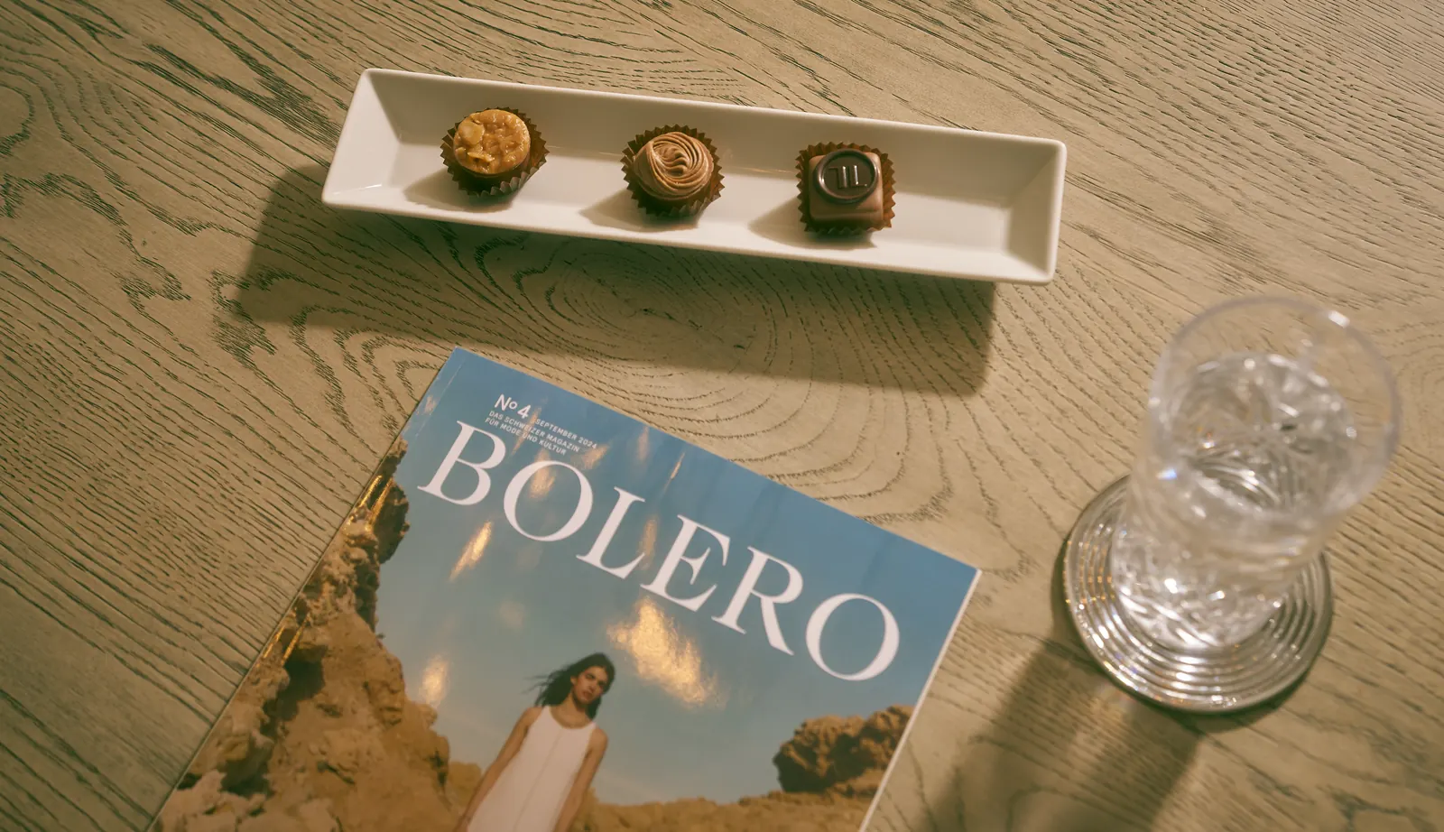 Three chocolates on a tray beside a magazine and a crystal glass of water on a wooden table