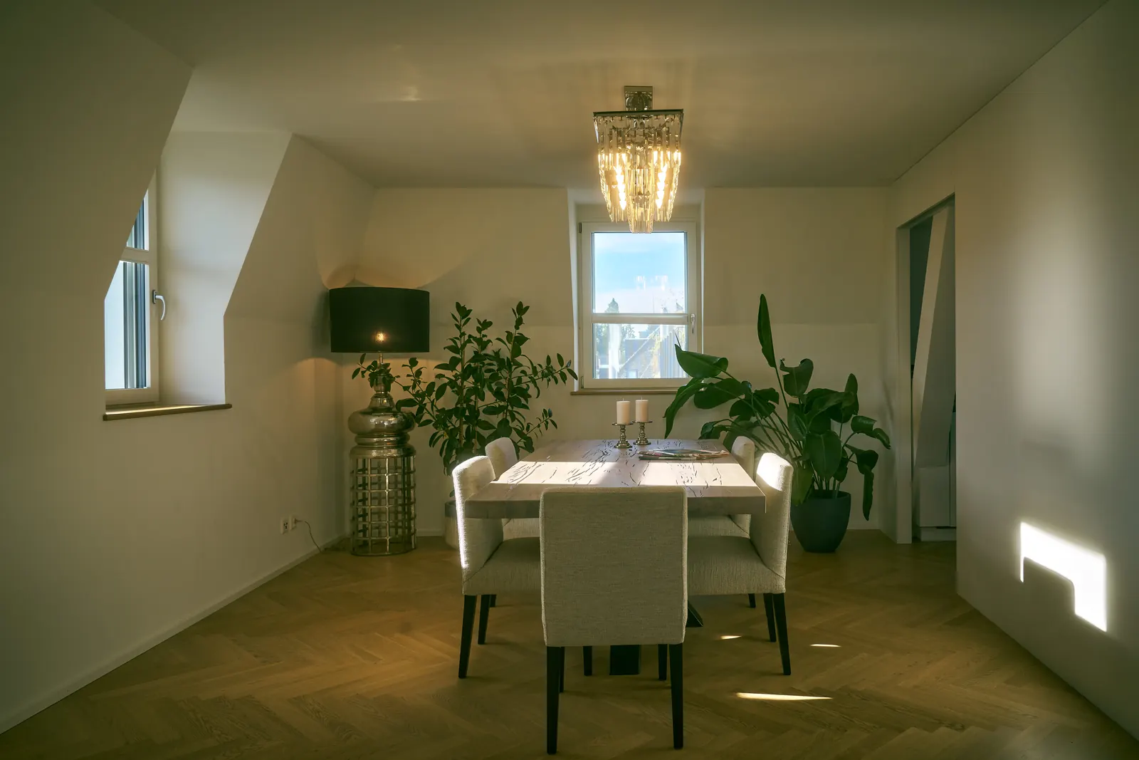 A dining table set with candles under a crystal chandelier, with plants and a window behind