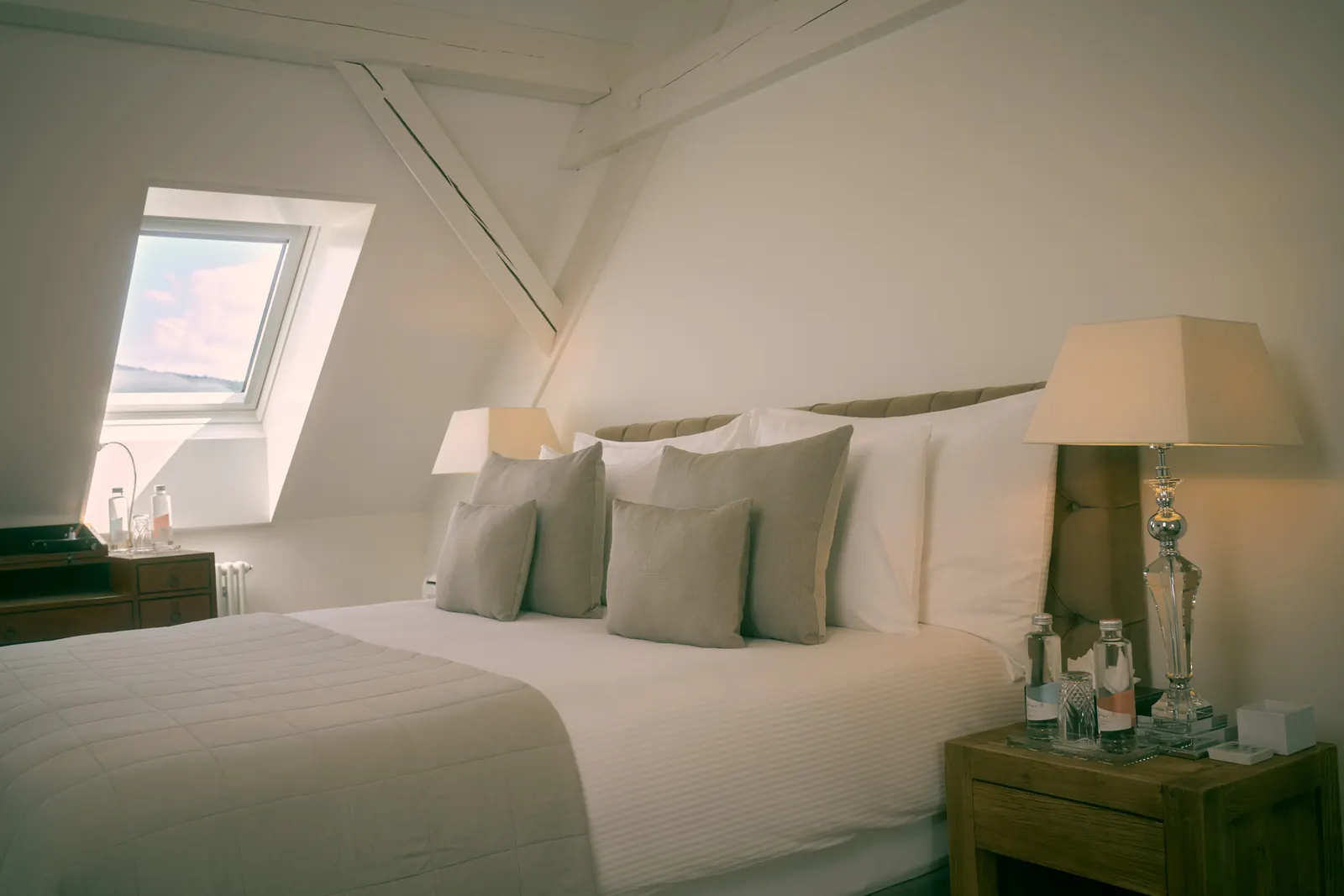 A bedroom under timber beams with twin skylights, a writing desk and a made bed