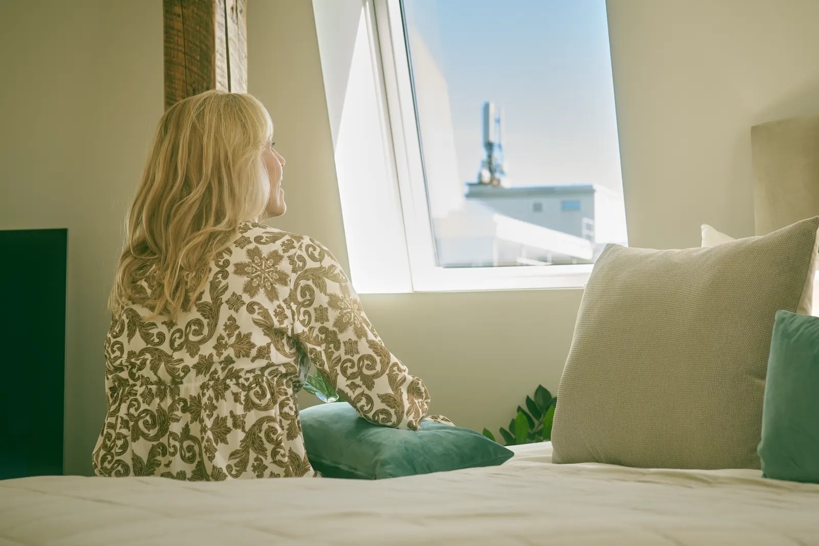 A person in a floral dress sits on a bed with their back to the camera, looking out of a window at the sky