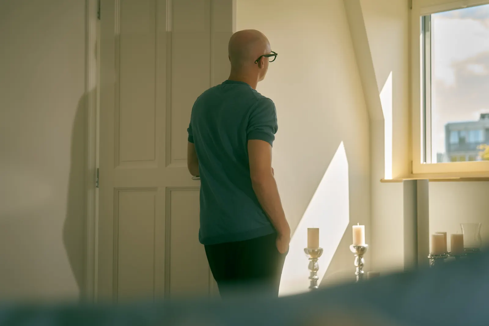 A person seen from behind stands at a sunlit window beside silver candlesticks