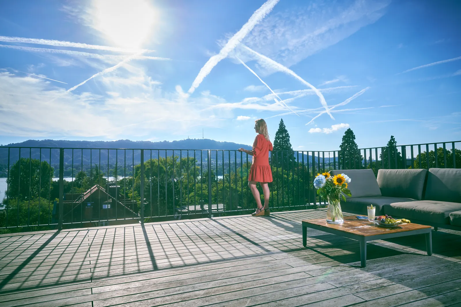 A person in a red dress, seen from a distance, stands at a roof terrace railing above Lake Zurich under a wide blue sky