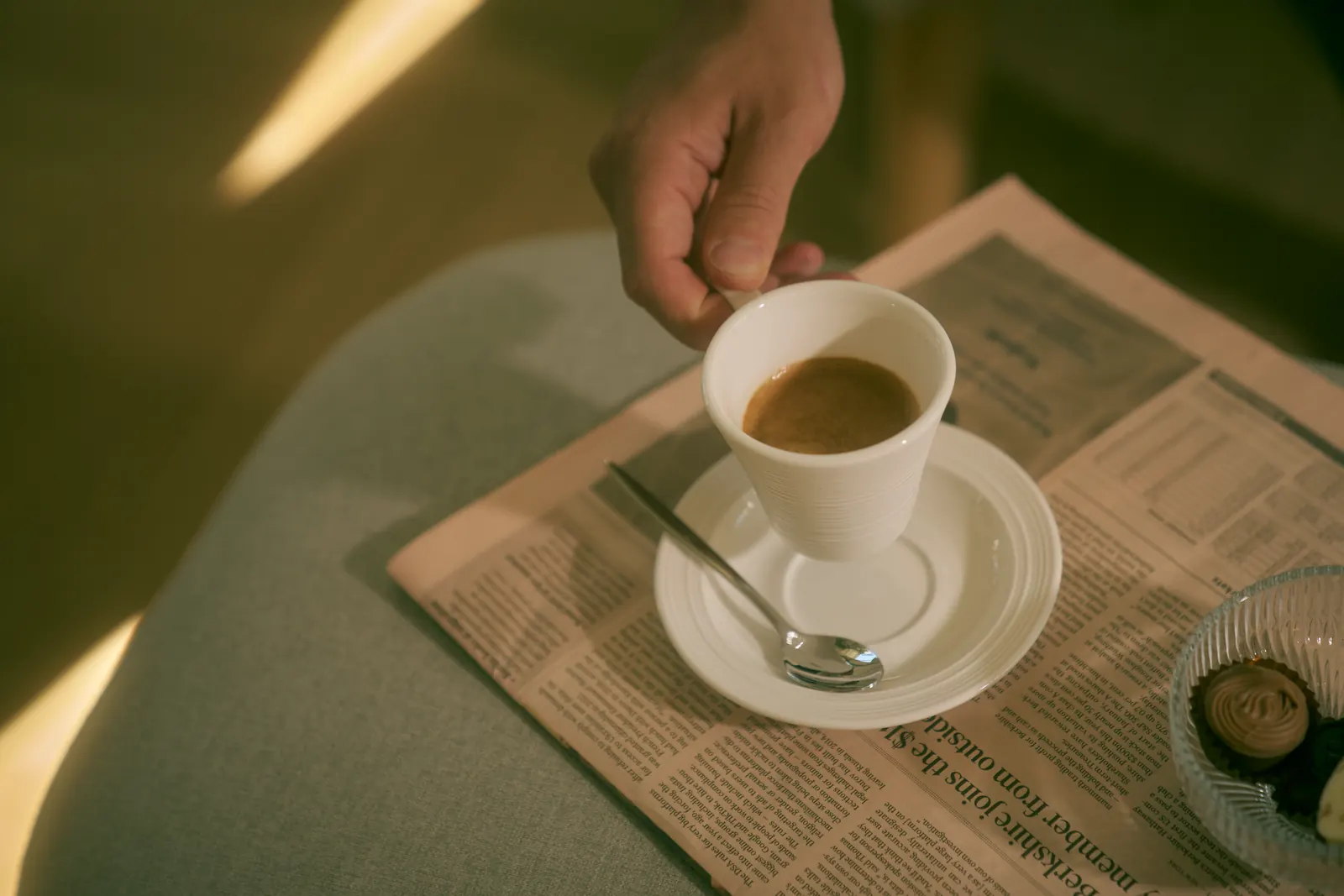 A hand lifts a small espresso cup from its saucer on a folded newspaper