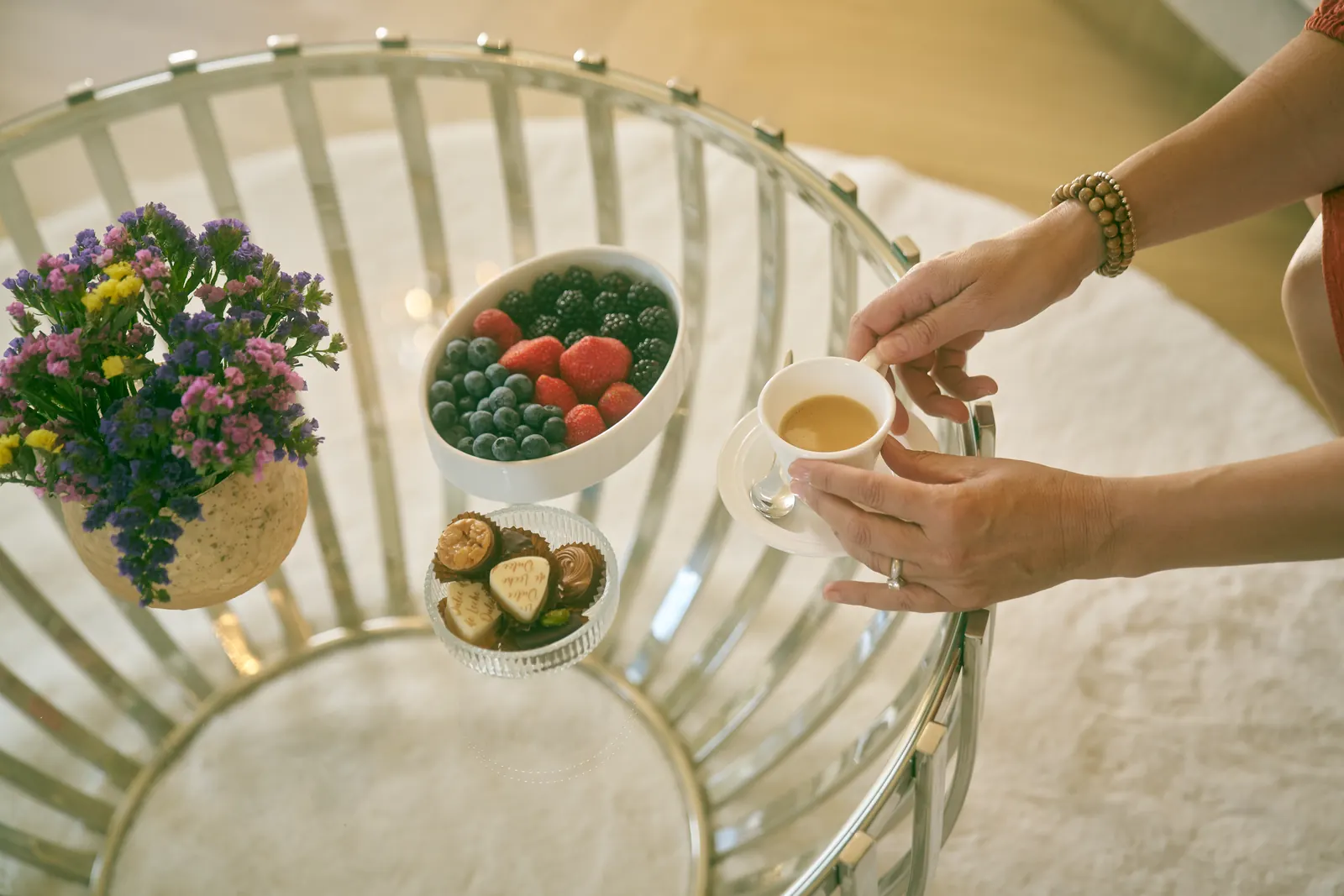 Hands lift a coffee cup from a glass table set with berries, chocolates and a small vase of flowers