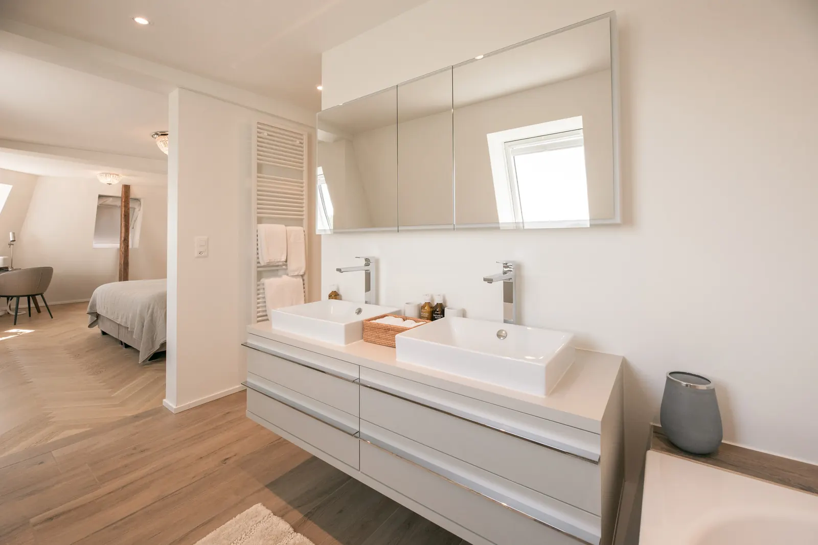 A bathroom with two basins and a mirrored cabinet, a bedroom visible through the doorway